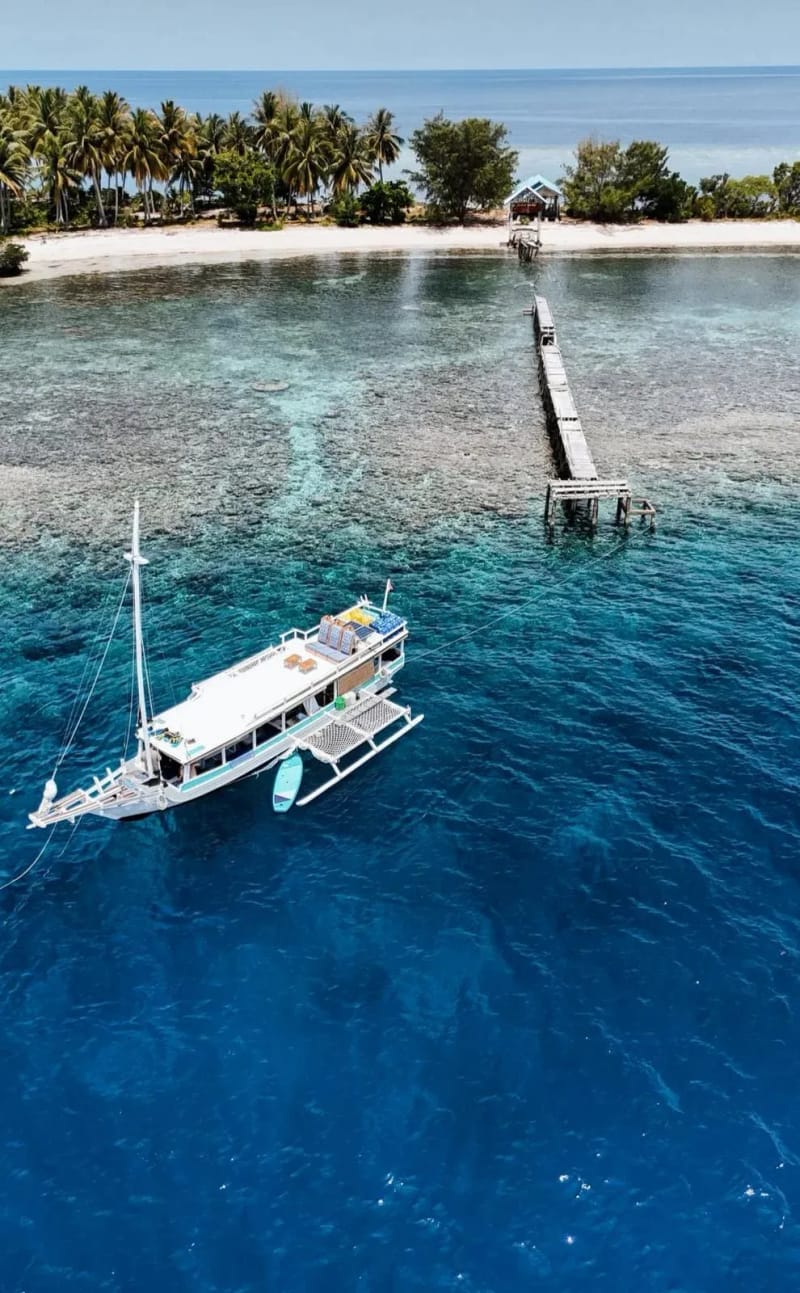 A phinisi anchored over a reef beside a long wooden jetty, seen from above