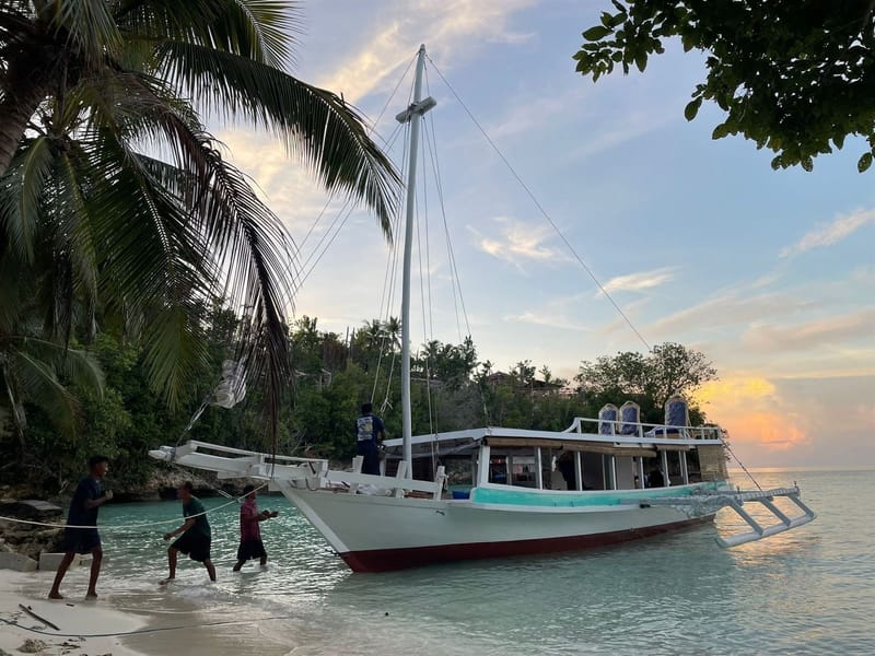 Crew bringing the white outrigger phinisi into a sandy beach under palm trees at sunset